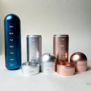 Könnte beinhalten: Eine Sammlung von Pillen-Organisatoren in verschiedenen metallischen Ausführungen. Die Organisatoren sind zylindrisch und mit Wochentagen beschriftet. Farben sind Blau, Silber und Roségold. Einige sind gestapelt, andere getrennt.