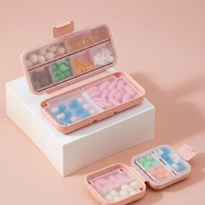 Puede incluir: Organizadores de pastillas rosas con múltiples compartimentos, llenos de varias pastillas y cápsulas de colores. Los organizadores están abiertos y cerrados, mostrando el contenido. El fondo es de un rosa suave, creando una estética limpia y organizada.