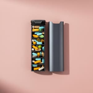 May include: A dark gray pill organizer with an open compartment filled with various colorful pills. The organizer is next to its matching lid, set against a soft pink background. The pills are in different shapes and colors.