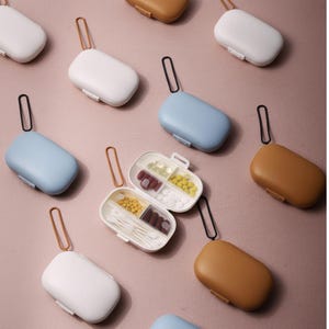 Puede incluir: Varios pastilleros rectangulares pequeños en colores variados, incluyendo blanco, azul y marrón. Cada estuche tiene un asa a juego y una tapa con bisagras. Uno está abierto, mostrando compartimentos llenos de pastillas y bastoncillos.