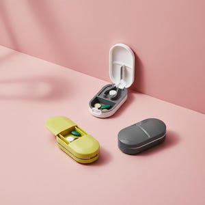 Könnte beinhalten: Drei Pillenboxen auf rosa Hintergrund. Eine ist offen, weiß und grau, mit Pillen darin. Eine andere ist gelb und offen, ebenfalls mit Pillen. Die dritte ist grau und geschlossen.