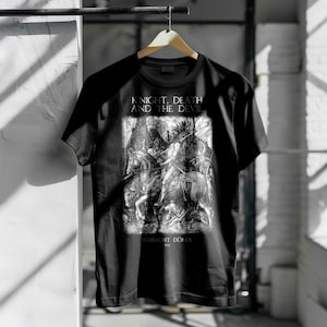 Puede incluir: Camiseta negra con una impresión en blanco y negro de la obra "El caballero, la muerte y el diablo" de Alberto Durero. El texto "KNIGHT, DEATH AND THE DEVIL" está impreso encima de la obra, con "ALBRECHT DÜRER" debajo.