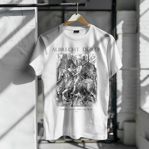 Puede incluir: Camiseta blanca con una impresión en blanco y negro de la obra "El caballero, la muerte y el diablo" de Albrecht Dürer. El texto "ALBRECHT DÜRER" está impreso encima de la obra, y "KNIGHT, DEATH AND THE DEVIL 1513" debajo.