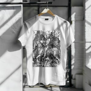 Puede incluir: Camiseta blanca con una ilustración en blanco y negro de una escena de batalla medieval. La imagen muestra caballeros a caballo, con detalles finos. La camiseta está colgada de una percha de madera.