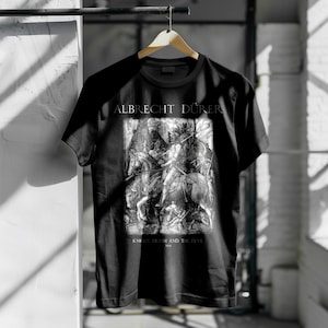 Op de afbeelding: Zwart t-shirt met een zwart-wit illustratie van een ridder te paard, met de tekst "ALBRECHT DÜRER" erboven en "KNIGHT, DEATH AND THE DEVIL" eronder. Het shirt hangt aan een houten hanger.