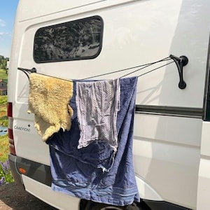 Op de afbeelding: Een zwart droogrek bevestigd aan de achterkant van een witte bestelwagen, met een beige schapenvacht, een grijs patroonhanddoek en een blauwe handdoek. De bestelwagen heeft een zwart raam en een muurschildering.