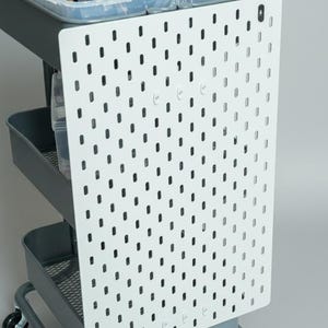 Peut inclure: Un panneau perforé blanc avec des trous espacés est fixé à un chariot roulant gris. Le chariot comporte trois bacs de rangement gris et une poignée. Le panneau perforé est conçu pour organiser les outils et accessoires.