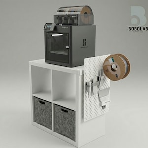 Op de afbeelding: Een grijze 3D-printer met een transparante filamentspoel erop, geplaatst op een witte opbergkast met grijze stoffen bakken. Een pegboard met accessoires en een filamentspoel is aan de zijkant bevestigd. Het logo "BOBOLAB" is zichtbaar.
