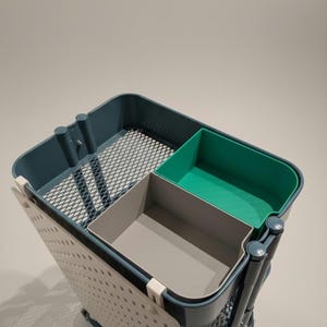 Op de afbeelding: Een rolwagen met een donkerblauw frame en een witte geperforeerde zijwand. De wagen heeft een metalen gaas bovenkant en bevat een grijze en een groene plastic opbergbak. Ontworpen voor organisatie.