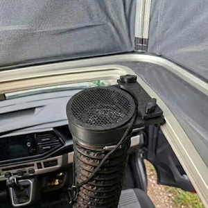 May include: Black air vent system with a mesh cover, mounted inside a vehicle. The vent is connected to a flexible, ribbed tube. The system is attached to the vehicle's frame with a black bracket. The interior of the vehicle is visible in the background.