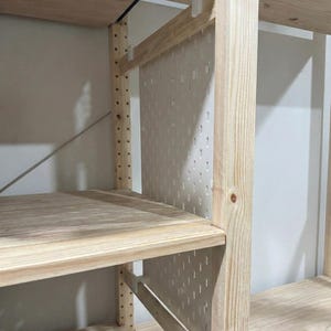 Könnte beinhalten: Regal aus Holz mit hellen Regalböden und Stützen. Das Regal hat eine Lochwand-Rückwand und weiße Regalstützen. Das Holz hat eine natürliche Oberfläche, und das Design ist einfach und funktional.