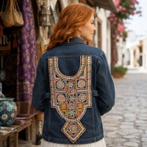 May include: A dark blue denim jacket featuring a large, intricate embroidered panel on the back. The embroidery includes colorful geometric patterns, circular motifs, and small reflective mirror accents arranged in a symmetrical, ornate design.