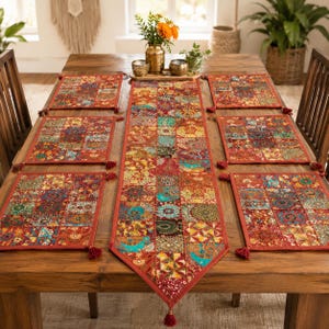 Puede incluir: Un camino de mesa rectangular y cuatro manteles individuales rectangulares a juego con un diseño de patchwork de varios patrones florales y geométricos coloridos en tonos rojo, naranja, verde y azul, acabados con pequeñas borlas rojas.