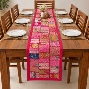 May include: A long, rectangular table runner featuring a vibrant patchwork design of various pink fabrics, intricate gold embroidery, sequins, and floral patterns, centered on a wooden dining table set with white plates and clear drinking glasses.
