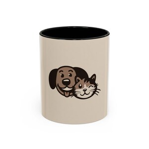 May include: A beige ceramic mug with a black interior. The mug features a cartoon illustration of a dog and a cat. The dog is brown with a black ear, and the cat is gray and white. The mug is suitable for hot or cold beverages.