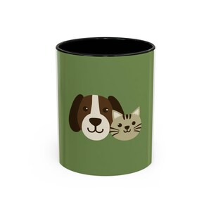 May include: A ceramic coffee mug with a green exterior and black interior. The mug features a cartoon illustration of a dog and a cat. The dog is brown and white, and the cat is grey. The mug is a standard 325 ml size.