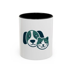 Peut inclure: Mug en céramique blanche avec un bord intérieur noir. Le mug présente un dessin bleu sarcelle et blanc d'un chien et d'un chat. Le chien a de grandes oreilles et le chat a une tache blanche sur le visage. Le mug est de taille standard.
