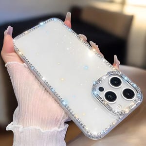 May include: A clear, white phone case with a sparkling glitter design and a border of small, clear rhinestones. The camera area is also outlined with rhinestones. The case is designed to fit a smartphone.