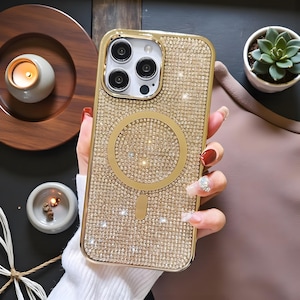 May include: Gold-colored phone case encrusted with small, sparkling rhinestones. The case has a gold frame and a circular design on the back. The phone case is shown with a gold-colored ring and a small, rectangular design at the bottom.