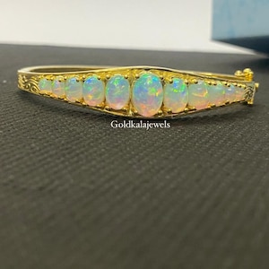 Puede incluir: Un brazalete rígido articulado de oro con una hilera de nueve ópalos ovalados iridiscentes dispuestos en degradado. La banda metálica presenta detalles de grabados intrincados en los laterales. El texto Goldkalajewels es visible.