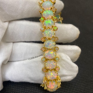 Puede incluir: Una pulsera de tono dorado que presenta una serie de ópalos redondos e iridiscentes engastados en garras de metal. Cada ópalo grande está flanqueado por ópalos de acento más pequeños, creando un diseño de eslabones. El texto Goldkalajewels está impreso.