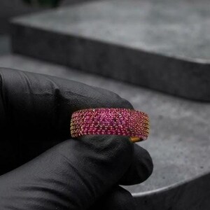 May include: A wide band ring featuring a dense pavé setting of numerous small, vibrant pink stones that cover the entire outer surface of the gold-toned metal band, creating a textured and uniform appearance across the circular jewelry piece.