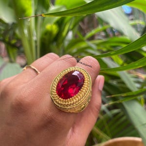 Puede incluir: Un anillo de oro con una gran piedra preciosa de color rojo intenso y corte ovalado, engastada en un borde texturizado tipo cuerda. La banda tiene un diseño ancho y ornamentado con detalles intrincados. El texto Goldkalajewels es visible.