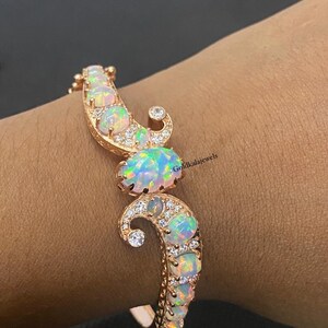 May include: A rose gold bracelet featuring a series of oval-shaped opals with iridescent flashes of color, accented by small clear stones set along the curved metal frame. The text Goldkalajewels is printed in small black letters on the metal surface.