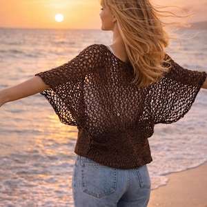 May include: A dark brown, short-sleeved, open-knit crochet top with a wide, loose fit and a solid-knit waistband, paired with light blue denim jeans. The top features a textured, net-like pattern across the shoulders and sleeves.