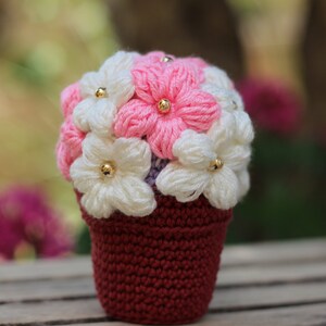 May include: A crocheted flower bouquet in a burgundy pot. The flowers are white and pink with gold centres.