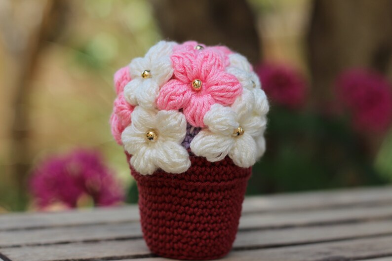 May include: A crocheted flower bouquet in a burgundy pot. The flowers are white and pink with gold centres.
