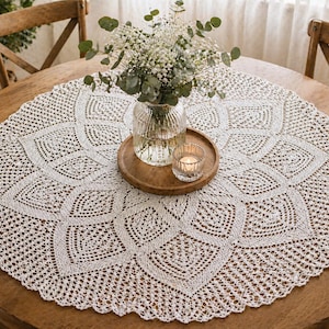 May include: A cream-coloured, circular lace tablecloth with a detailed floral pattern is displayed on a wooden table. A glass vase with white flowers, a wooden tray, and a lit candle are centered. Two wooden chairs are visible in the background.