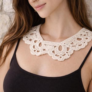 May include: A cream-colored, crocheted collar with a scalloped edge and openwork design. The collar is worn over a black tank top. The image is a close-up of the collar and upper body.