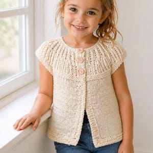 Könnte beinhalten: Eine cremefarbene Strickweste mit Knopfleiste und strukturiertem Design. Die Weste hat einen runden Ausschnitt und drei rosa Knöpfe. Das Kind trägt blaue Jeans.