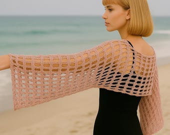 May include: A light pink, open-weave shrug with long sleeves. The shrug is made of a soft, knitted material and has a loose, flowing fit. The garment is worn over a black top. The background is a beach with the ocean in the distance.