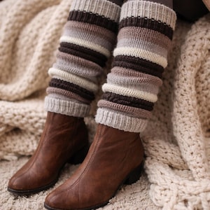 Puede incluir: Calentadores de piernas de punto en tonos marrón, gris y crema, usados sobre leggings grises y combinados con botas de cuero marrones. Los calentadores de piernas presentan rayas horizontales. Las botas tienen un tacón bajo y están sobre una alfombra crema.