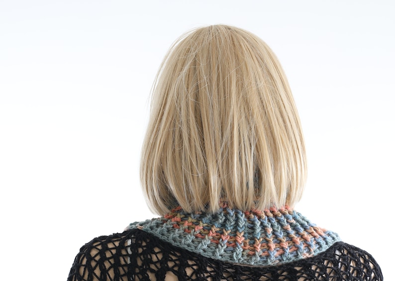 May include: A woman with blonde hair is wearing a crocheted neck warmer with a multi-colored pattern of blue, green, and orange. The neck warmer is made of a soft, chunky yarn.