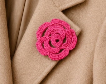 May include: A vibrant fuchsia crochet flower pin is attached to a tan wool coat. The handmade floral accessory adds a pop of color and texture. The flower is a detailed, three-dimensional design, perfect for adding a touch of elegance to any outfit.