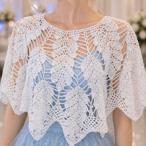 May include: A white, open-weave crochet shawl with a floral pattern. The shawl is worn over a light blue dress. The crochet work is detailed, creating a delicate and airy appearance. The style is suitable for special occasions.