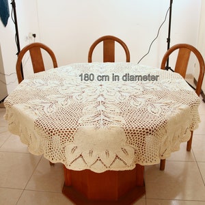 Puede incluir: Un mantel de crochet blanco con un delicado patr&oacute;n floral, colocado sobre una mesa de madera con cuatro sillas. El mantel tiene un di&aacute;metro de 180 cm.