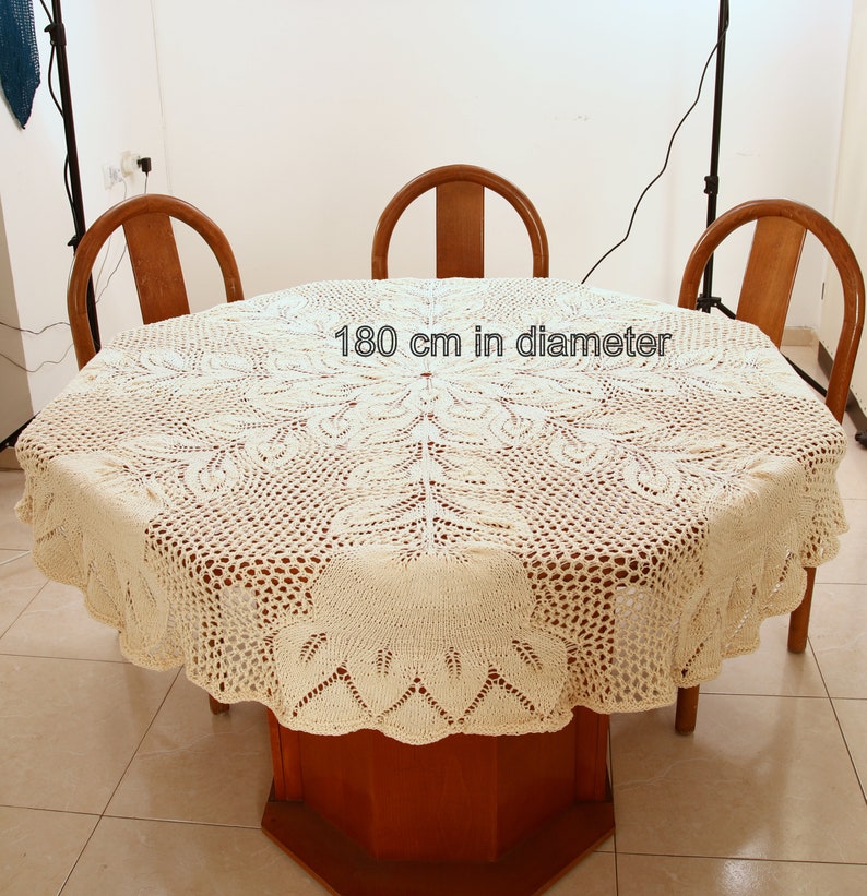 Puede incluir: Un mantel de crochet blanco con un delicado patr&oacute;n floral, colocado sobre una mesa de madera con cuatro sillas. El mantel tiene un di&aacute;metro de 180 cm.