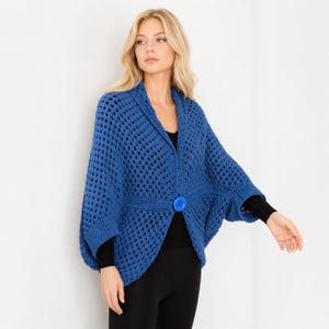 Pode incluir: Um cardigan azul de crochê com fecho de botão grande. O design de tecido aberto do cardigan é visível. As mangas são compridas e a gola é estilo xale. O cardigan é usado sobre um top preto e leggings pretas.