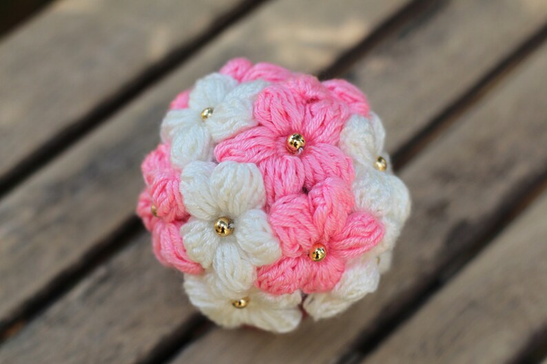 May include: A crocheted flower bouquet made of pink and white yarn with gold beads in the centre of each flower.
