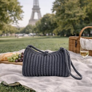 May include: A dark gray, crocheted handbag with a ribbed design. The bag has a rounded shape with a handle and a button closure. The background includes a picnic setting with a basket, food, and the Eiffel Tower.