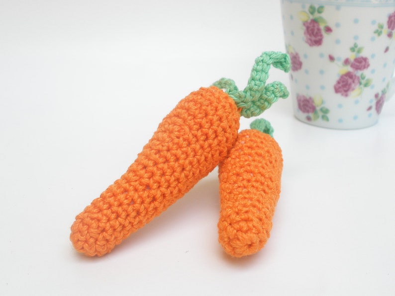 May include: Two crocheted carrots, one orange with green leaves, the other orange. The carrots are on a white surface.