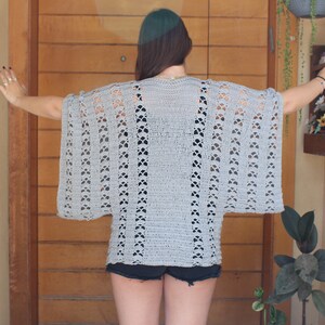 May include: A light grey, open-weave crochet top with wide sleeves. The top features a repeating floral pattern along the sleeves and vertical lines. The garment is worn over a dark top and shorts.