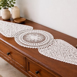 May include: A cream lace table runner with a central circular motif and two pointed ends, displayed on a dark wooden table. The runner is made of open-weave fabric. The table also features vases and a framed picture.