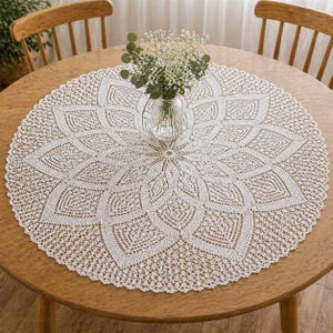 May include: A circular, white, crocheted tablecloth with a floral pattern on a wooden table. A clear glass vase with a small bouquet of white flowers is in the centre. Two wooden chairs are in the background. The table is made of wood.