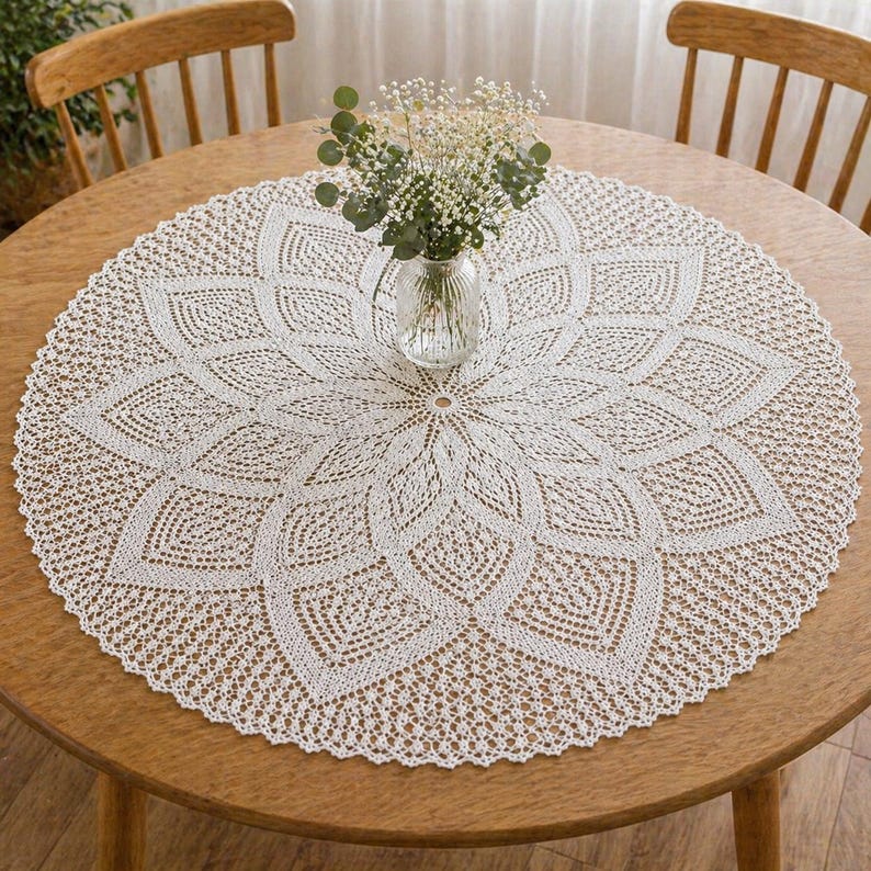 May include: A circular, white, crocheted tablecloth with a floral pattern on a wooden table. A clear glass vase with a small bouquet of white flowers is in the centre. Two wooden chairs are in the background. The table is made of wood.