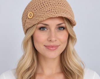 May include: A tan crocheted newsboy cap featuring a textured, open-weave pattern and a short, stiff brim. A single, round, light-colored wooden button is attached to the side of the cap, providing a decorative accent to the structured design.